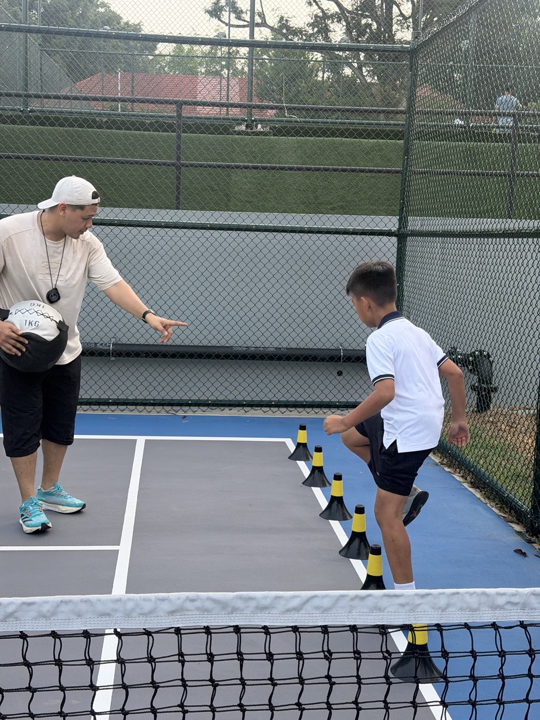A coach in a white cap holding a 1 kg medicine ball points at a line of yellow-and-black agility cones as a young athlete steps through them on an outdoor court.