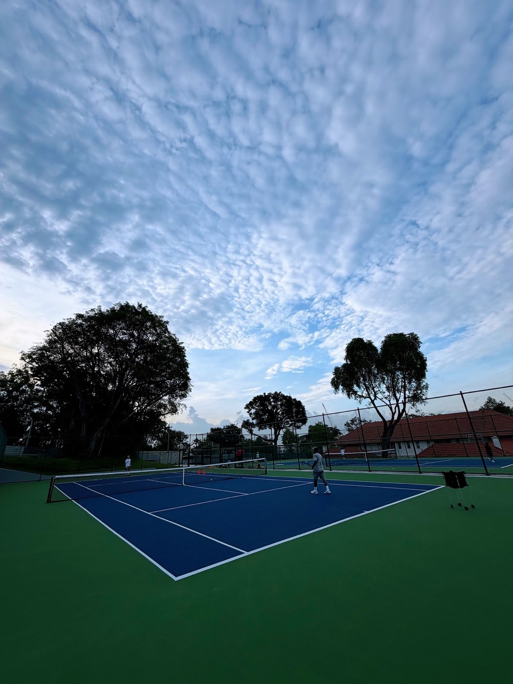 A player on an outdoor hard court at dusk, with training cones and a wheeled ball cart nearby.
