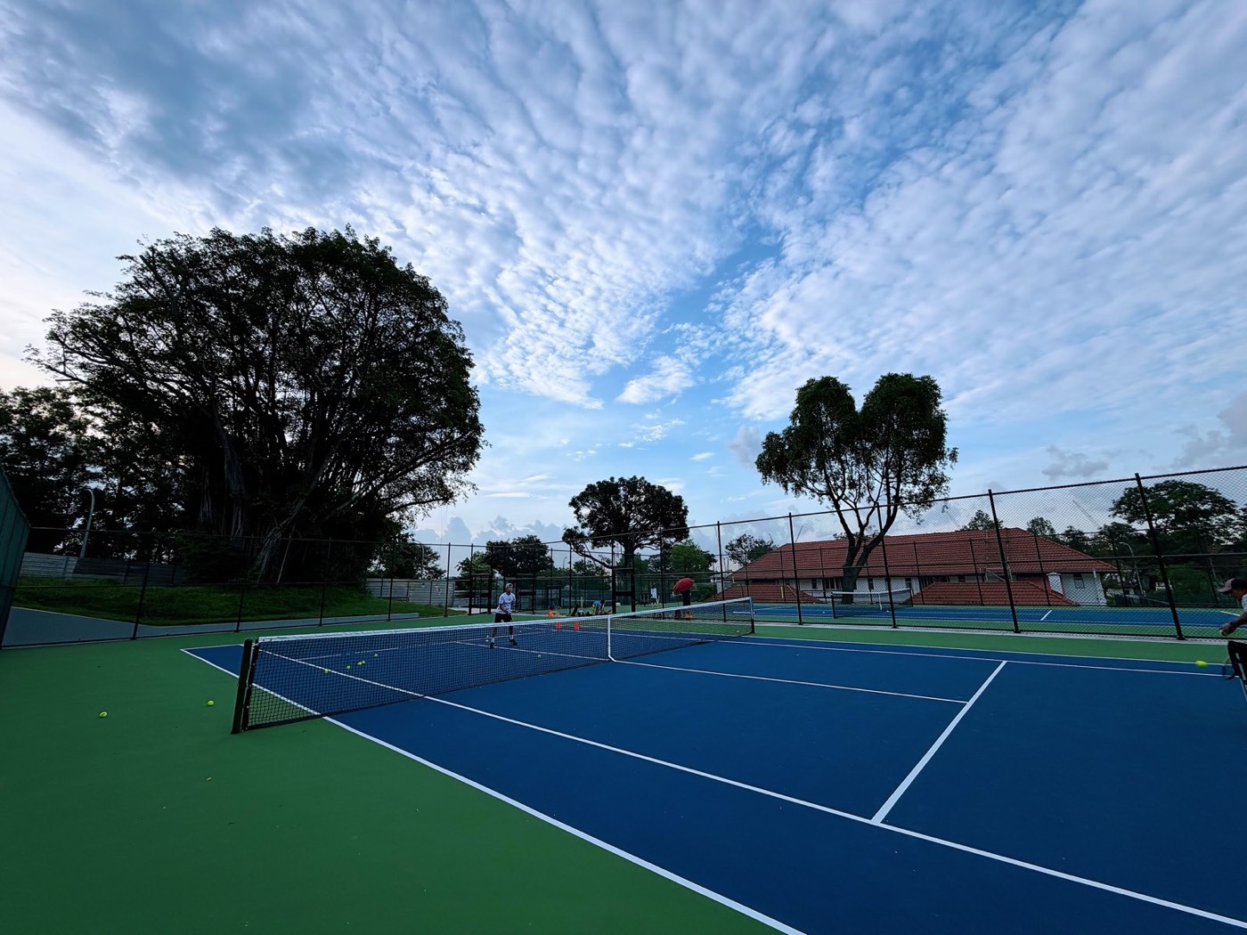 Outdoor hard courts at dusk with tennis balls and orange training cones on court.
