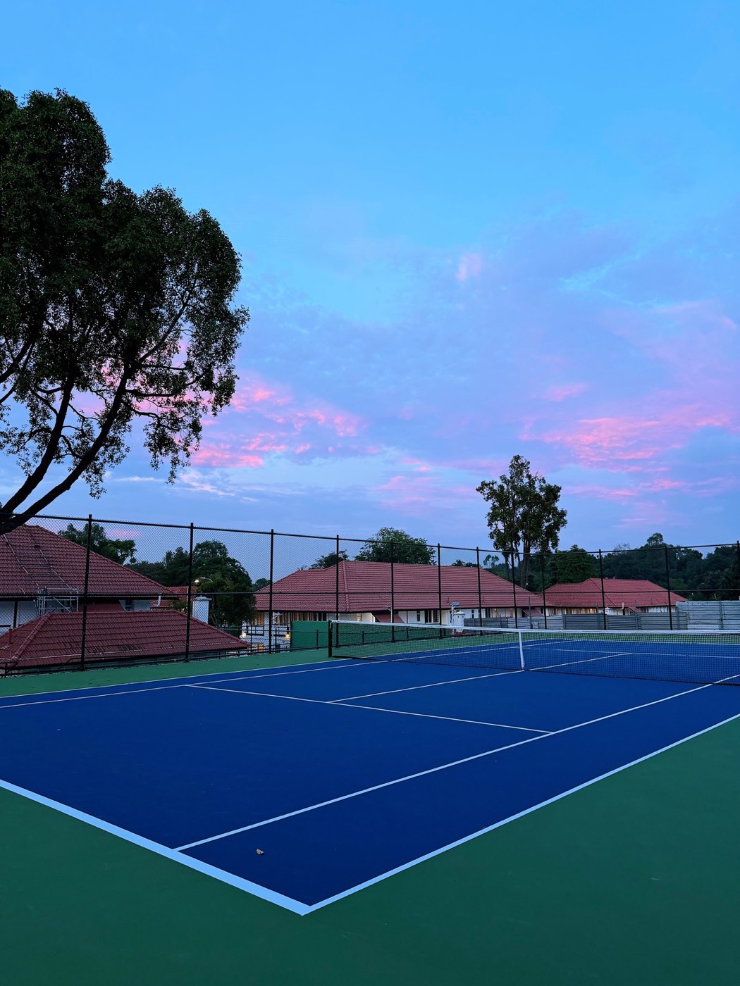 An empty outdoor hard tennis court at dusk behind a chain-link fence, with tile-roofed buildings beyond.