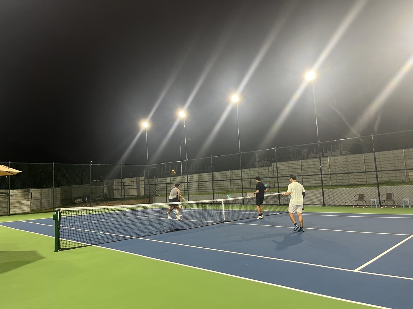 Three adults with rackets near the net on a floodlit outdoor tennis court at night.