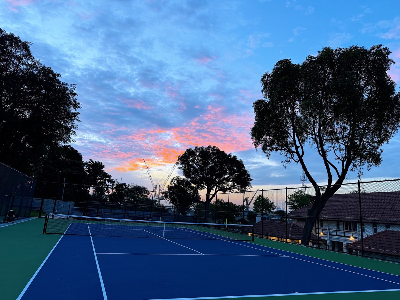 An empty outdoor tennis court with a blue playing surface at sunset, framed by silhouetted trees.