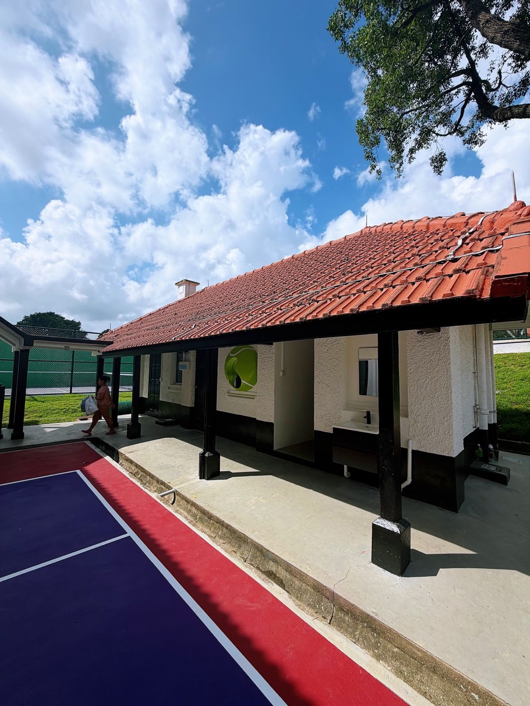 A single-storey pavilion with a red tile roof and open veranda beside a small hard court.